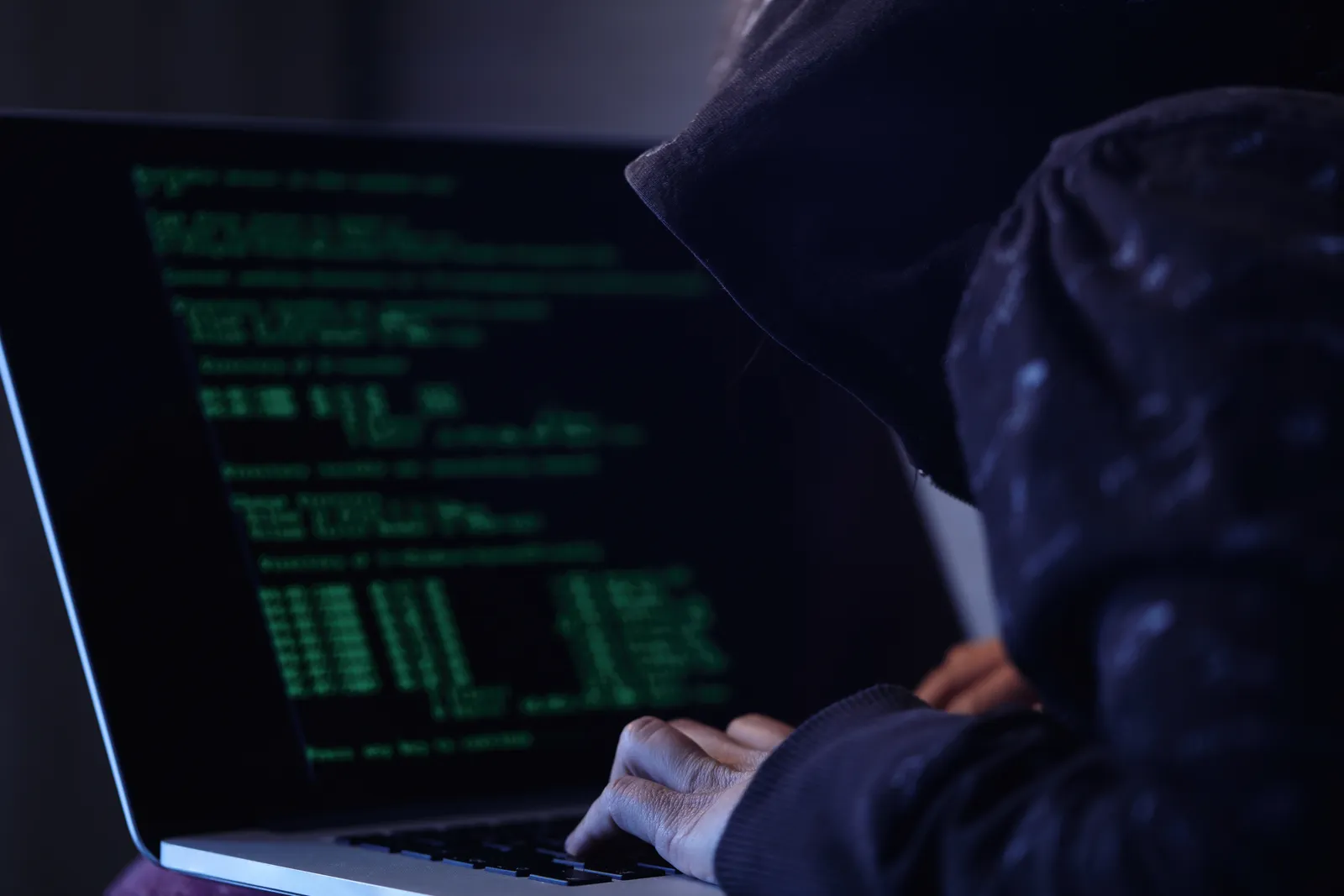 Person im Kapuzenpulli tippt an einem Laptop mit grünem Programmcode